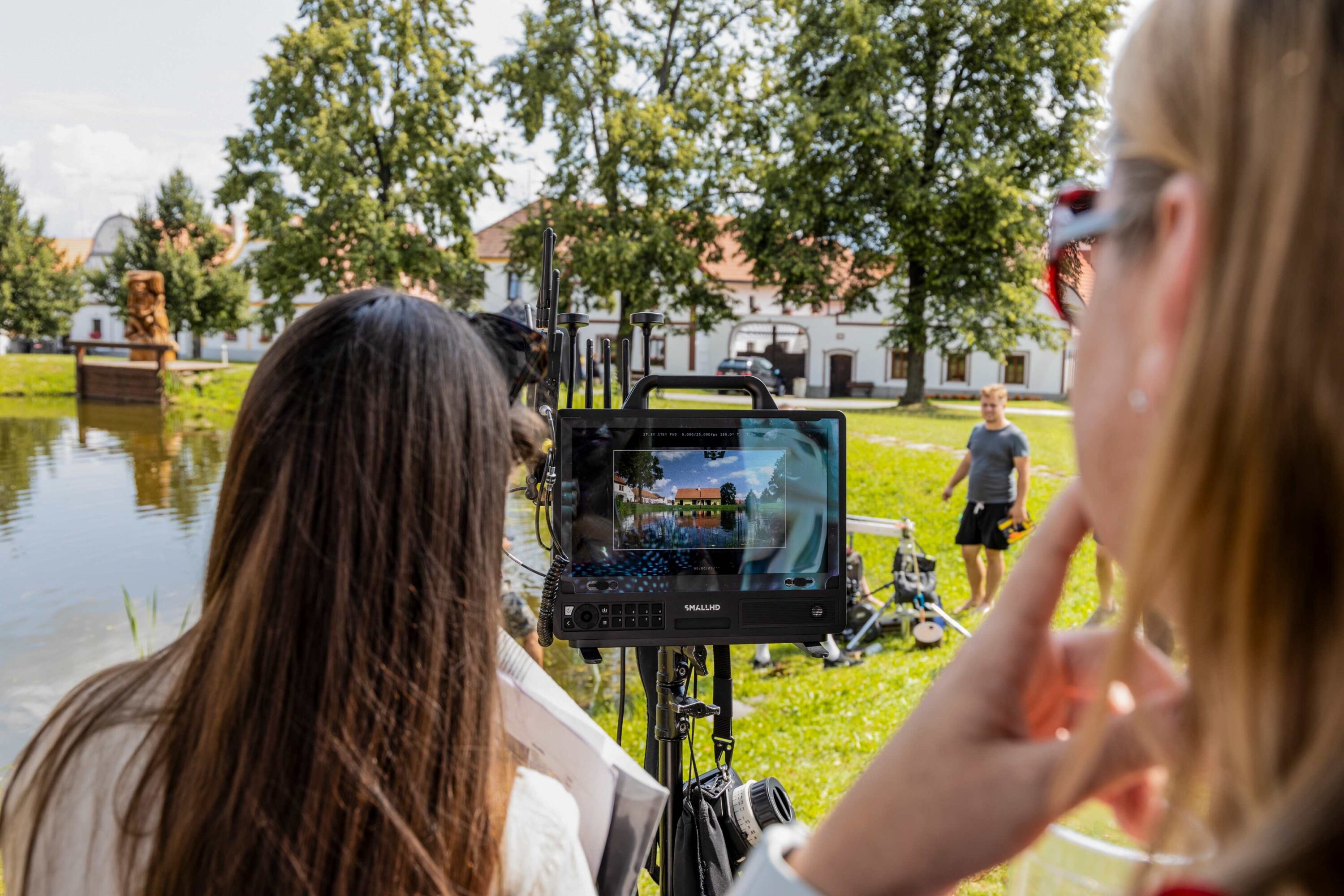 Crew outdoors filming scene using a monitor on a rig beside a lake and trees behind them, with people in foreground and background.