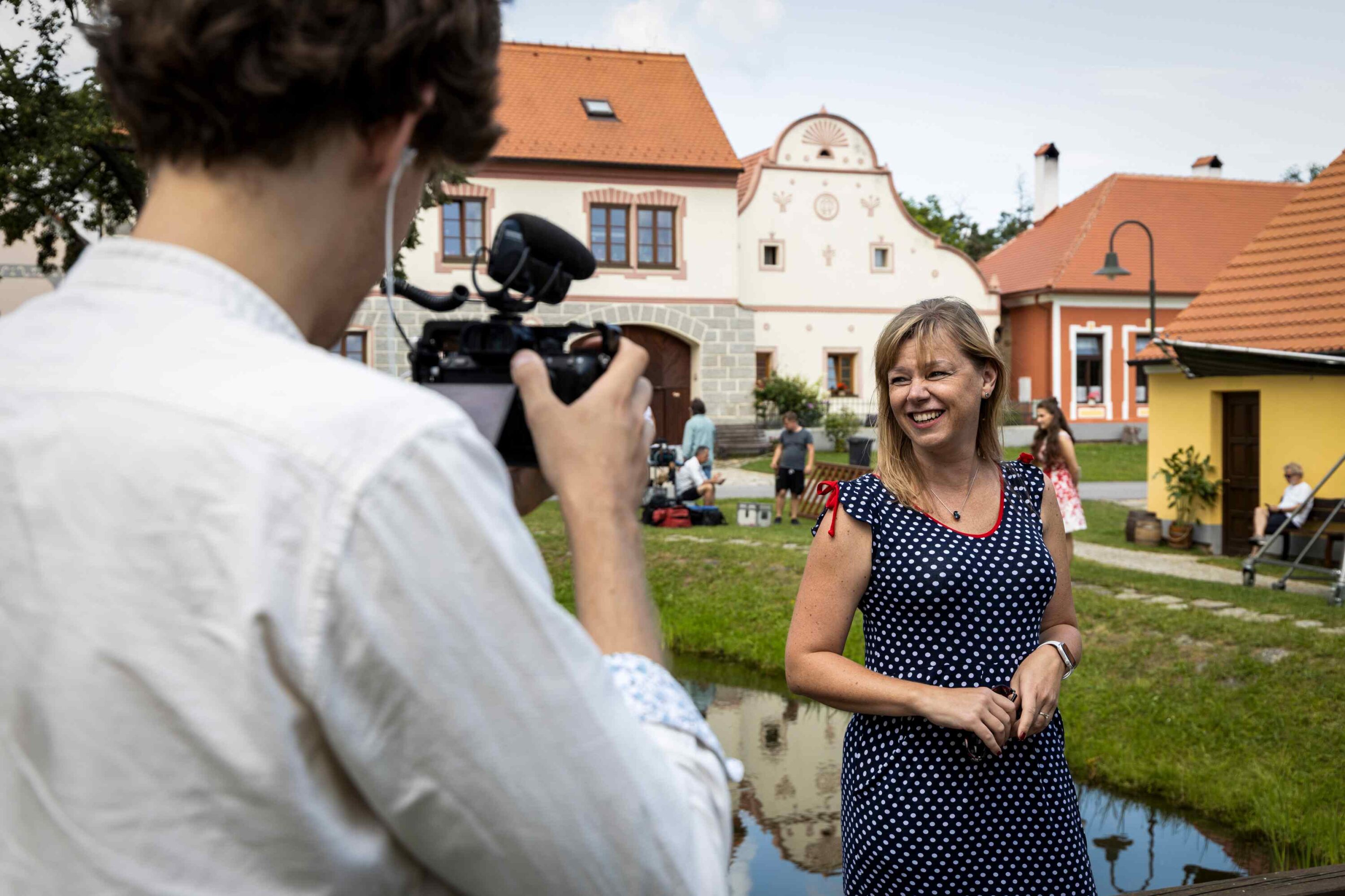 Cameraman filming a smiling woman in a navy polka-dot dress outdoors near colorful town buildings.
