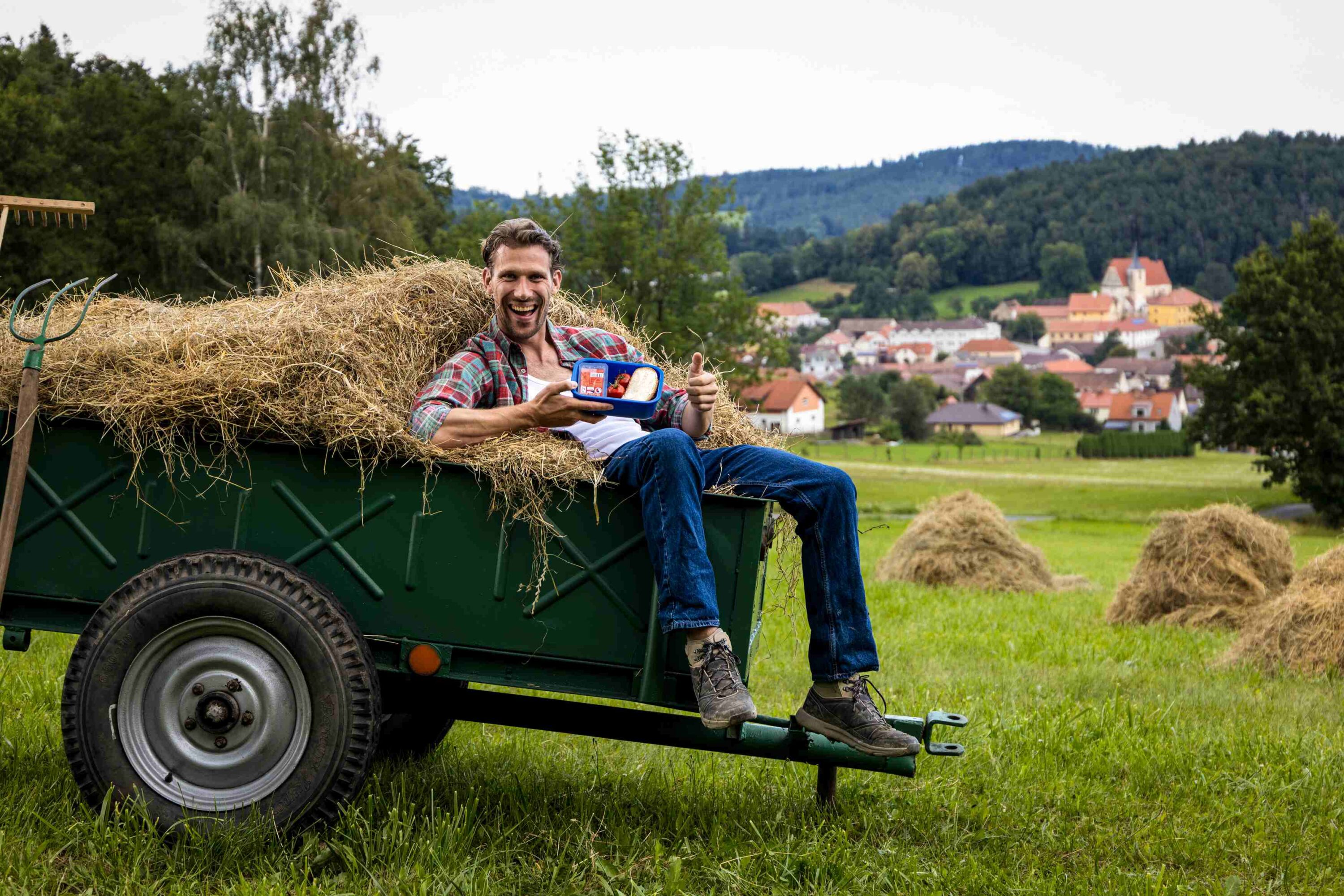 Man sitting on a hay-filled trailer in a field, smiling and giving a thumbs-up with a small food container in hand; a village and hills in the background.