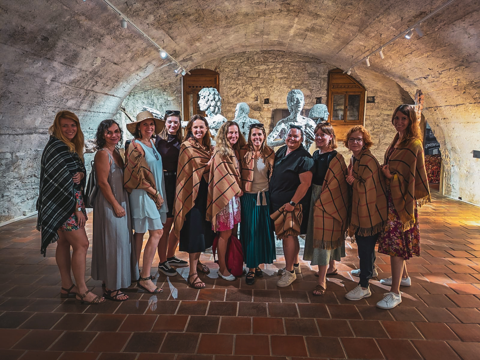 Group of women posing together in a stone-walled gallery, many wearing tan plaid shawls, in a historic exhibit space.
