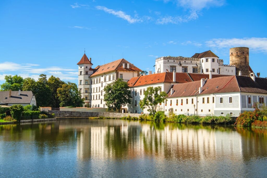 Historic castle with white walls and red-tiled roofs along a calm river, reflecting in the water under a blue sky.