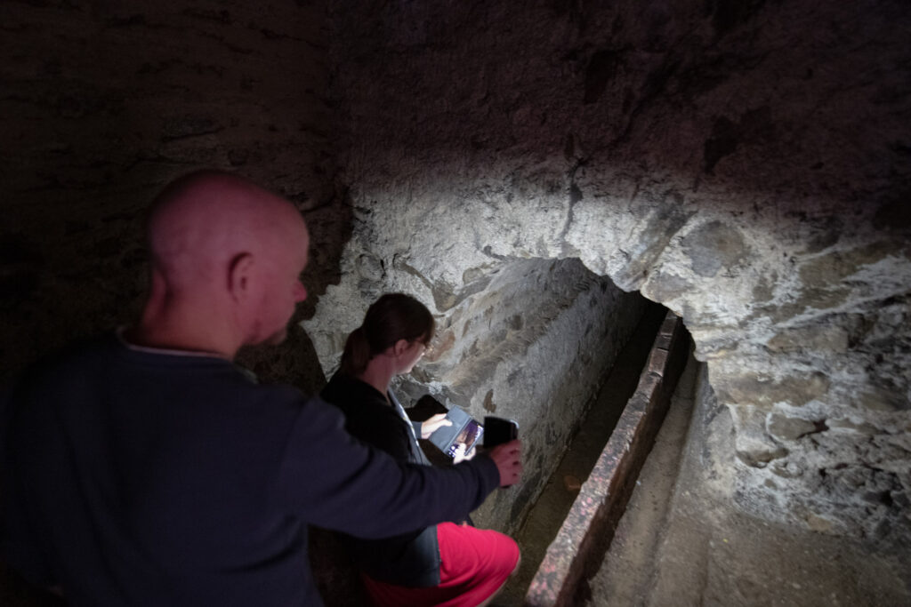 Účastníci Location Tour fotí v úzké kamenné chodbě slavonického podzemí s odvodňovacím kanálem
