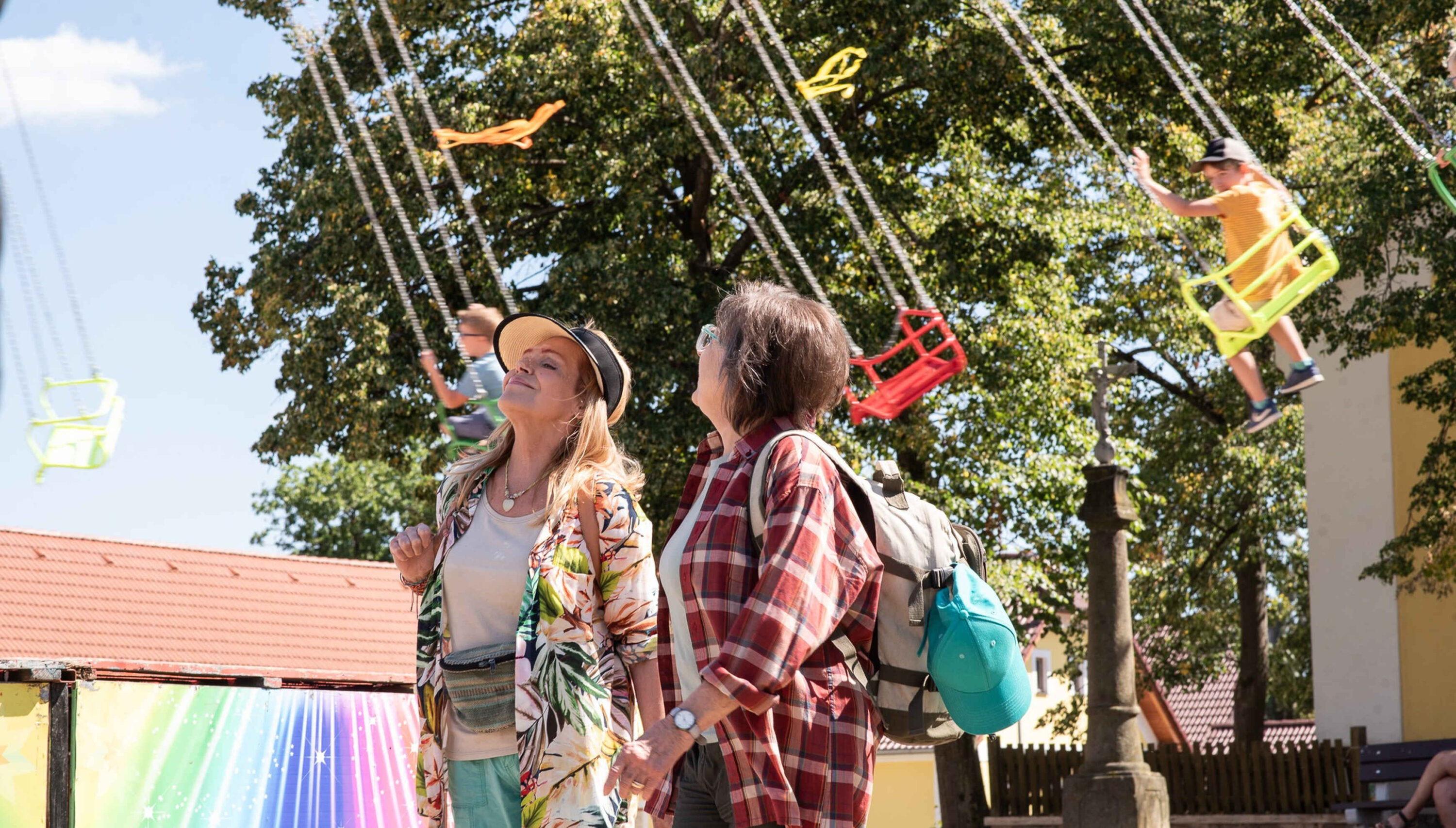 Two adults walking together and smiling as a child on a bright yellow swing rides overhead on a sunny day in a park.