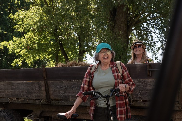 Two smiling women outdoors near a wooden hay wagon; one holds a bike and wears a teal cap and plaid shirt, with a second woman in the background wearing a hat.