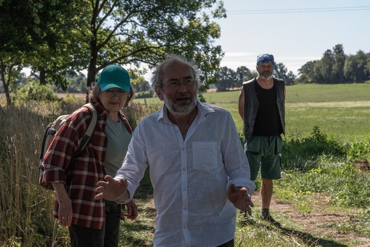 Man in a white shirt speaks and gestures toward the camera as two companions stand behind in a sunny rural field.