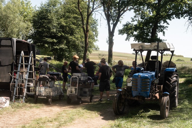 Group of people outdoors in a rural yard, standing around equipment and carts near a blue tractor under trees.