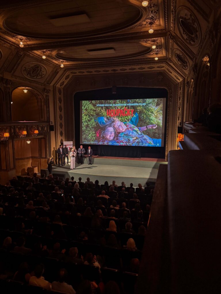 Luxurious ornate theater interior with a large stage screen showing a colorful movie poster and a group of people standing onstage, audience in the dark.