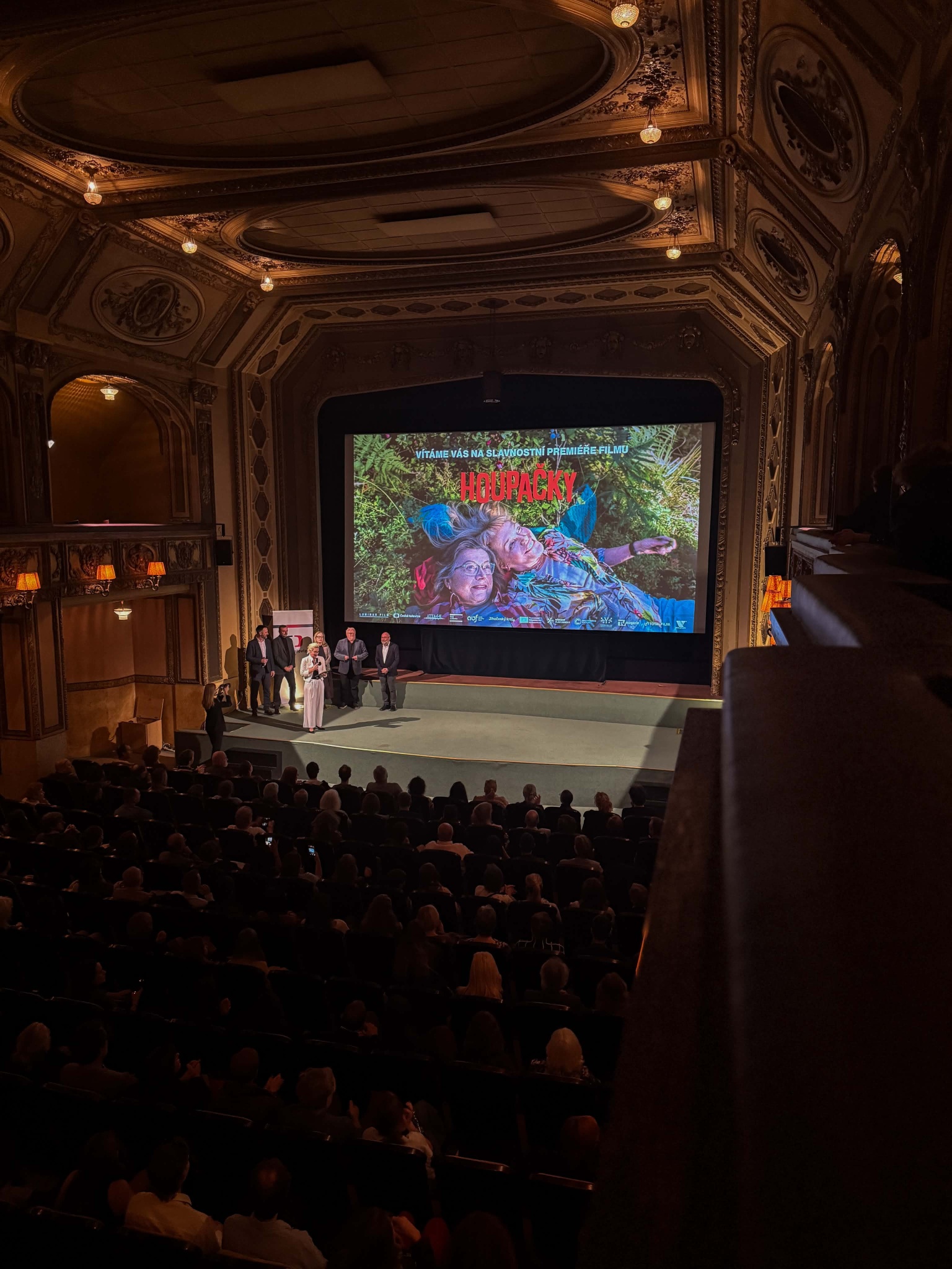 Luxurious ornate theater interior with a large stage screen showing a colorful movie poster and a group of people standing onstage, audience in the dark.